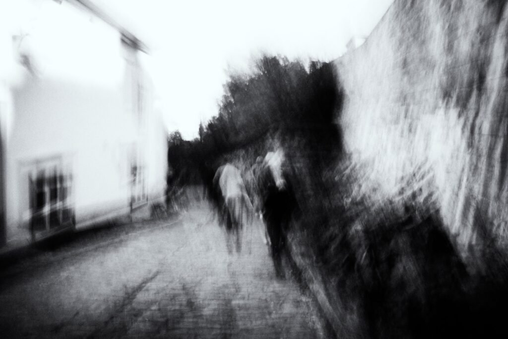 Chemin bordé d’un mur, groupe de silhouettes floues avançant, mouvement diagonal en noir et blanc.