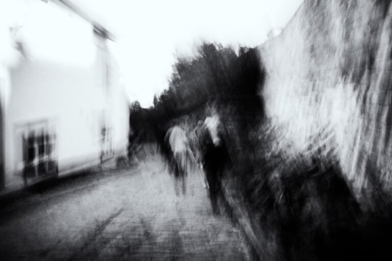 Chemin bordé d’un mur, groupe de silhouettes floues avançant, mouvement diagonal en noir et blanc.