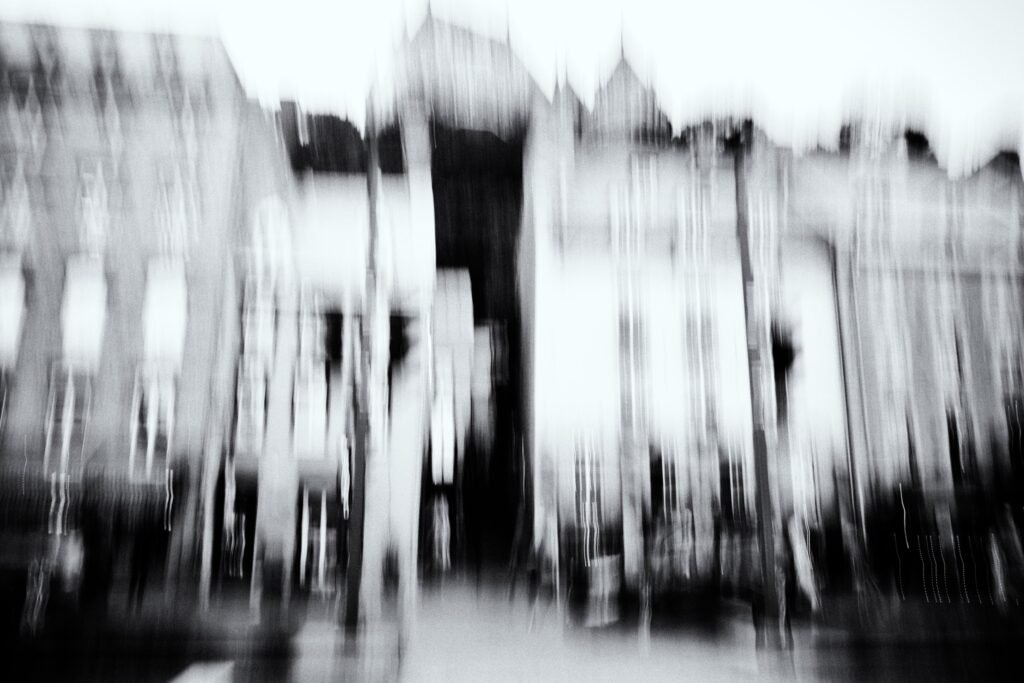 Architecture en verticales, silhouettes fines, flou directionnel, noir et blanc, détails partiellement effacés.