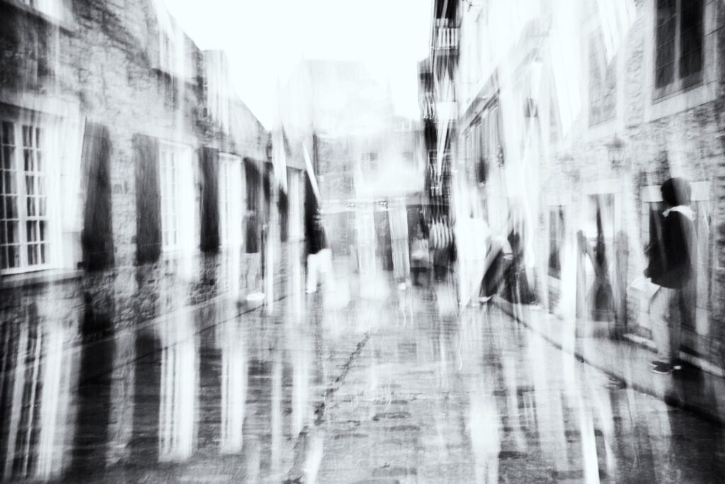 Rue pavée humide, reflets au sol, silhouettes floues, lignes de fuite en noir et blanc.